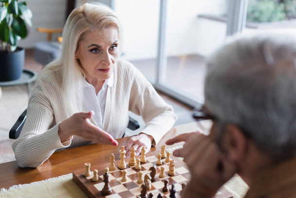 Seniorin zeigt auf Schach neben verschwommenem Ehemann zu Hause  - Foto, Bild