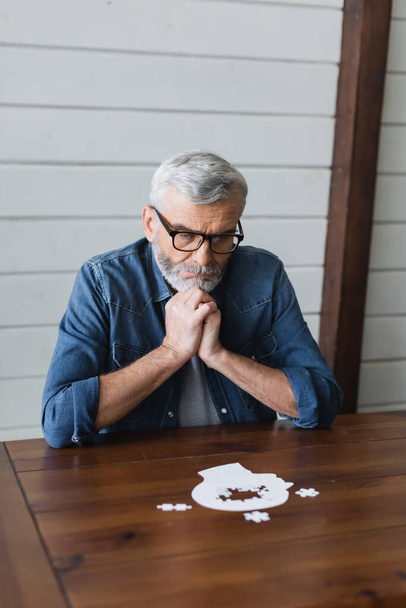 Figyelmes idős férfi szemüvegben, homályos kirakóst bámul az asztalon.   - Fotó, kép