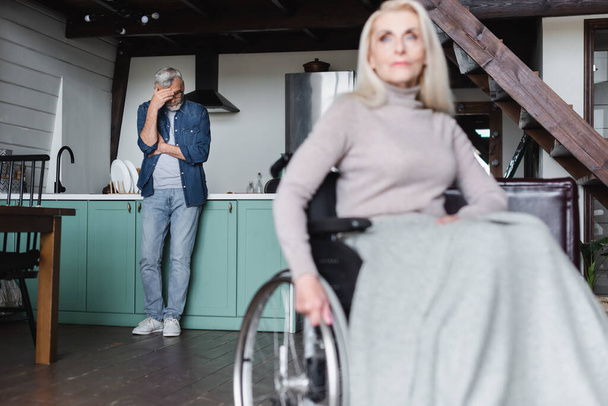 Θλιβερός άνθρωπος στέκεται στην κουζίνα κοντά θολή γυναίκα σε αναπηρικό καροτσάκι  - Φωτογραφία, εικόνα