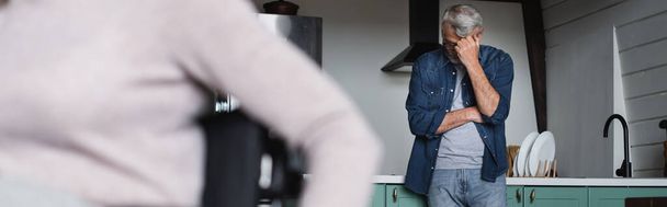 自宅でぼやけた妻の近くのキッチンに立つ動揺シニア男、バナー  - 写真・画像