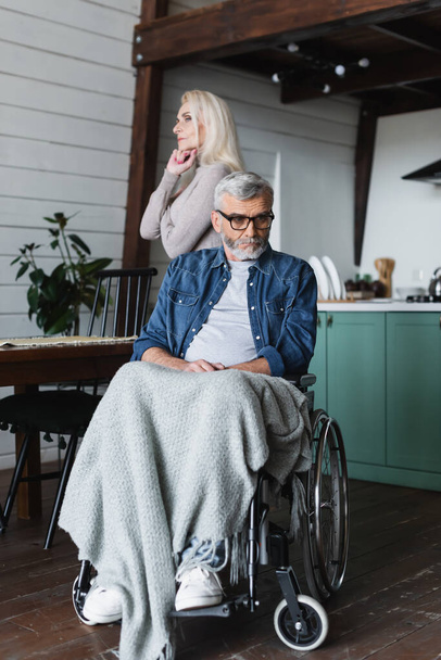 Λυπημένος ανάπηρος άνδρας σε αναπηρική καρέκλα κοιτάζει μακριά κοντά στη γυναίκα του στο σπίτι  - Φωτογραφία, εικόνα