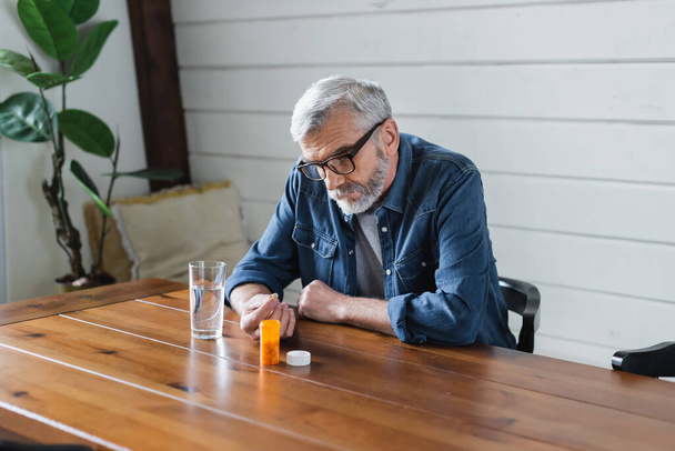 Lonely senior man holding pill near water on table  - Fotografie, Obrázek