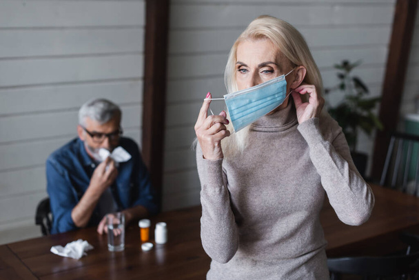 Женщина в медицинской маске рядом с размытым мужем с салфеткой и таблетками  - Фото, изображение
