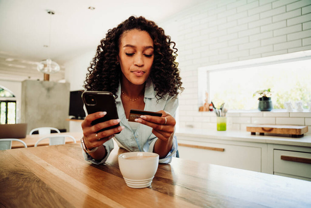 Mixed race female making online payment standing in kitchen with credit card and cellphone - Foto, Bild