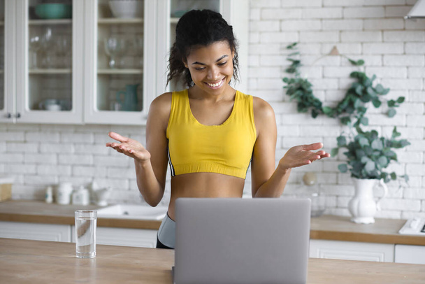 Young beautiful African American woman fitness trainer using laptop by video call, provides an online consultation on healthy eating and fitness standing in the home kitchen - 写真・画像