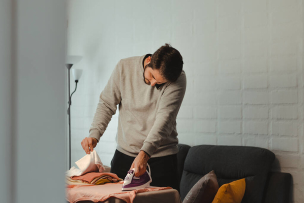 Hombre latino planchando ropa de mujer en la sala de estar mientras habla por teléfono celular. Adulto joven haciendo tareas domésticas. Concepto de igualdad de género. - Foto, imagen