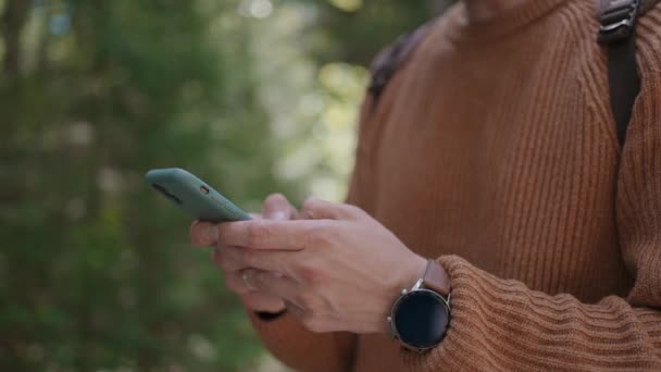 sorrindo homem com óculos com barba caminha pela floresta com uma mochila e imprime uma mensagem em seu telefone celular em câmera lenta. - Filmagem, Vídeo