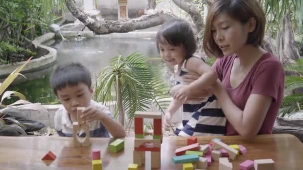楽しい、幸せな男の子と女の子の学習スキルで創造と開発のためのパズル木製ブロックおもちゃをプレイする若いアジアの母親と子供たち自宅でゲームで活動するためのデスクで、教育の概念. - 映像、動画