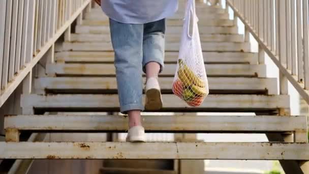Frau in Jeans geht die Treppe hinauf und hält weiße Netztasche mit Früchten in der Hand. Umweltfreundliche, wiederverwendbare Einkaufstasche. Orangen, Äpfel, Bananen in Baumwoll-Stricktasche. Null Abfall und plastikfreies Konzept. - Filmmaterial, Video