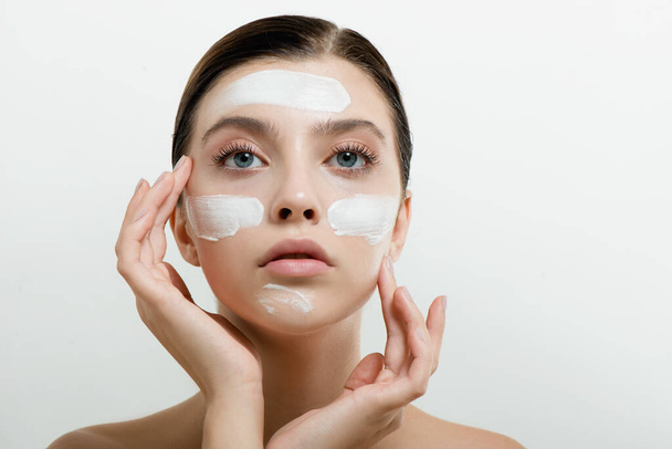 Young woman applying face cream or facial mask at her face. Beauty model with perfect fresh skin and long eyelashes cares about her skin at home. Spa and Wellness, Skin Care Concept. Close up, selected focus. - Photo, image