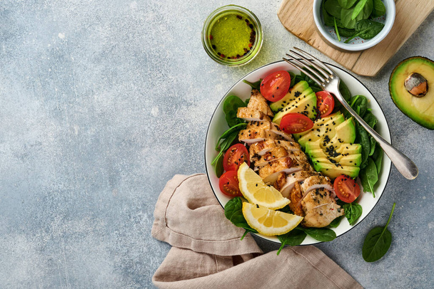 Fresh green salad with grilled chicken fillet, spinach, tomatoes, avocado, lemon and black sesame seeds with olive oil in white bowl on light slate background. Nutrition Diet Concept. Top view. - Foto, Bild
