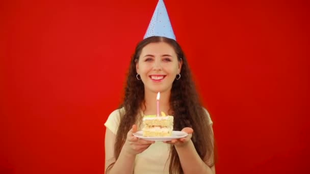 Νεαρή γυναίκα σε εορταστικό καπέλο κρατά ένα πιάτο με μια φέτα κέικ και ένα κερί απομονωμένο σε κόκκινο φόντο. - Πλάνα, βίντεο