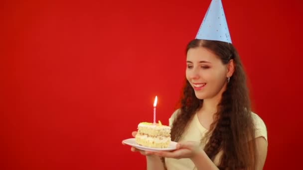 Tentative infructueuse d'éteindre la bougie et de faire un vœu d'anniversaire. Jeune femme dans une casquette festive tient une assiette avec une tranche de gâteau et une bougie allumée isolée sur un fond rouge. - Séquence, vidéo