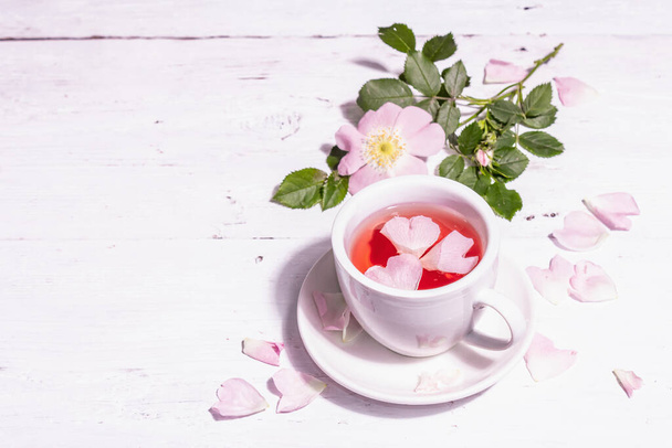 Herbata z kwiatów różanych bioder. Letni napój witaminowy, mocne światło, ciemny cień. Białe drewniane tło, przestrzeń do kopiowania - Zdjęcie, obraz