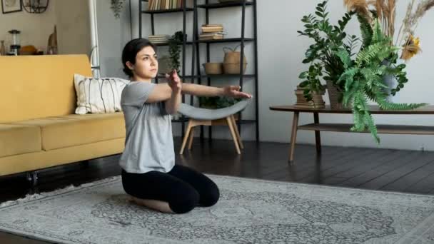 Una donna indiana si siede sul pavimento e si prepara per un allenamento di yoga, fa un riscaldamento e stretching dei muscoli, un esercizio di torsione delle braccia. Situato a casa in una camera accogliente - Filmati, video