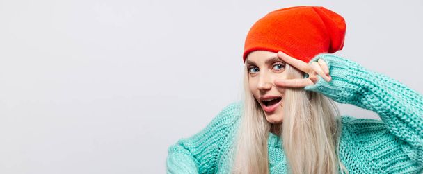 Studio portrait of happy young blonde girl showing peace sign on white background. Wearing orange hat and blue sweater. - Фото, изображение