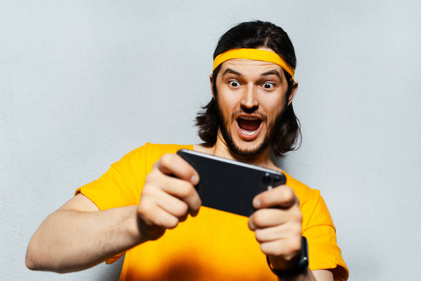 Studio muotokuva nuori yllättynyt mies pitkät hiukset, pelaa peliä älypuhelimella harmaa kuvioitu tausta; yllään keltainen bändi pään ja paidan. - Valokuva, kuva