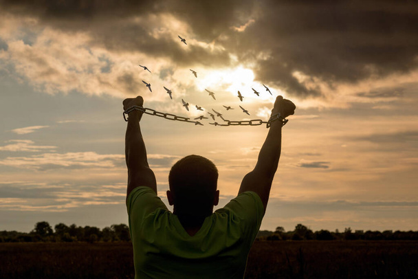 Un jeune homme brise la chaîne qui s'effrite en oiseaux sur fond de coucher de soleil ensoleillé. - Photo, image