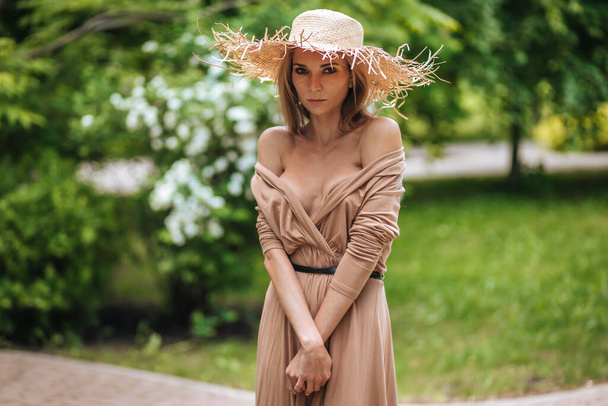 Retrato de una hermosa mujer sexy en un vestido con un escote y un sombrero de paja en un parque de verano - Foto, Imagen