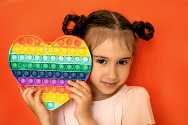 Симпатичная девушка с косичками в руках с большой сердечной игрушкой. Студийный портрет ребенка на красном фоне. Выразительные красивые карие глаза - Фото, изображение