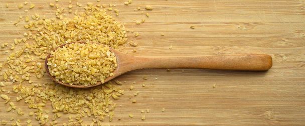 Dry cracked wheat daliya grains in wooden spoon, on bamboo cutting board - Photo, Image