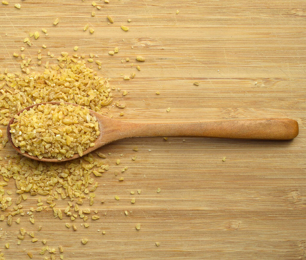 Dry cracked wheat daliya grains in wooden spoon, on bamboo cutting board - Fotó, kép