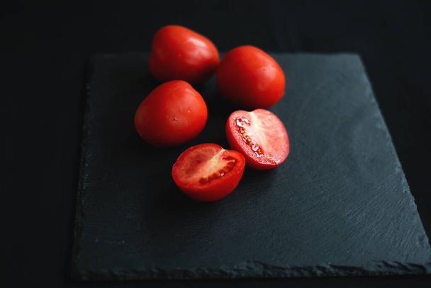 skupina rajčat, jedna rozpůlená, spočívá na černé desce pro servírování - Fotografie, Obrázek