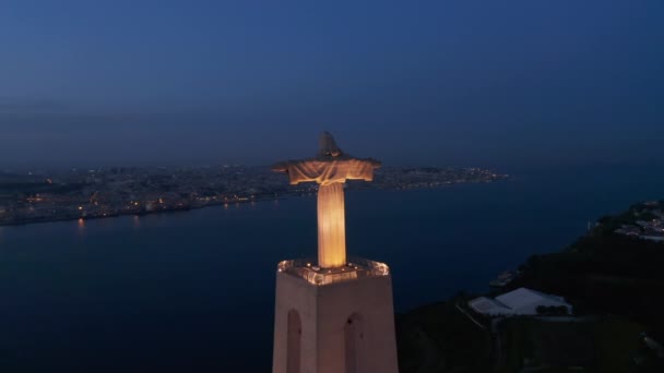 Νύχτα drone πετούν προς μεγάλο φωτισμένο άγαλμα του Ιησού με απλωμένα χέρια. Αεροφωτογραφία του Ιερού του Βασιλιά Χριστού στην Αλμάδα μετά το ηλιοβασίλεμα. Λισαβόνα, πρωτεύουσα της Πορτογαλίας. - Πλάνα, βίντεο
