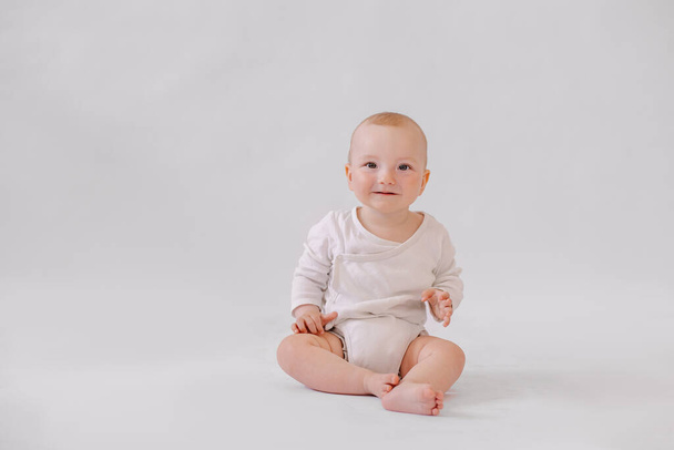 cute little kid in a white bodysuit sits on  background. 8 months old boy smiling.  - Φωτογραφία, εικόνα