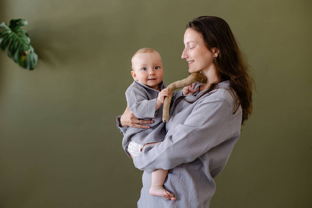 mother in grey clothes pajamas hug a baby.  Mom and child. Happiness of motherhood. Eco parenting vegan. Closeness to nature. Happy childhood. Natural fabric. Green background copyspace. lifestyle kid - Photo, image