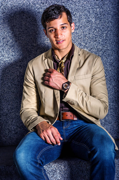 Young Man Fashion. Ein junger Mann in beigem Blazer, blauer Jeans, einem kleinen Schal und einer Armbanduhr sitzt da und schaut einen an. - Foto, Bild