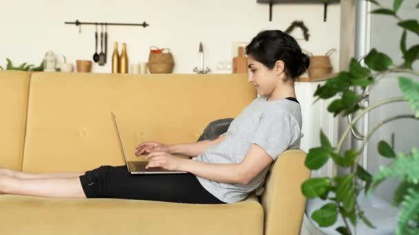 Woman using laptop at home, A Girl Is Lying on a Yellow Sofa in a Bright Room. Businesswoman working on laptop at remote workplace. Work from home concept. Remote learning concept. - Filmagem, Vídeo