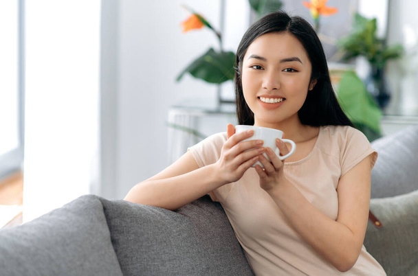 Портрет счастливой красивой азиатской брюнетки, сидящей на диване в гостиной, одетой в повседневную стильную одежду, пьющей чай или кофе, смотрящей в камеру, расслабляющей, улыбающейся - Фото, изображение