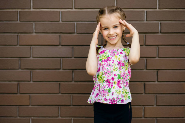 Krásná veselá holčička se usměje a drží si ruce u hlavy. Malá holčička v letním jasném oblečení stojí u cihlové zdi a usmívá se roztomile. Volné místo pro text - Fotografie, Obrázek