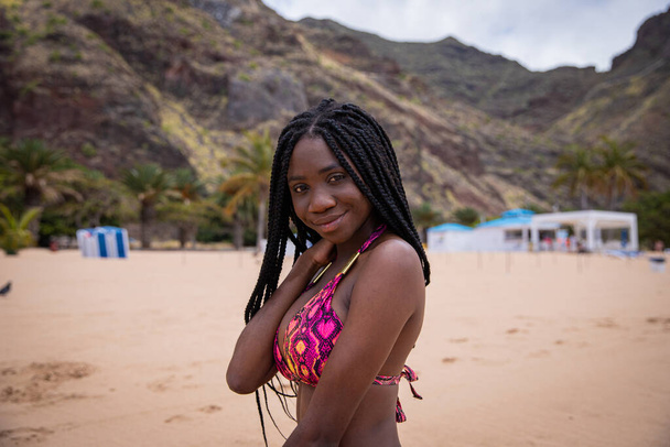 Porträt eines schönen lächelnden schwarzen Mädchens am Strand. Sie trägt einen Bikini und hat Zöpfe, sie ist unbeschwert und genießt den Urlaub. - Foto, Bild