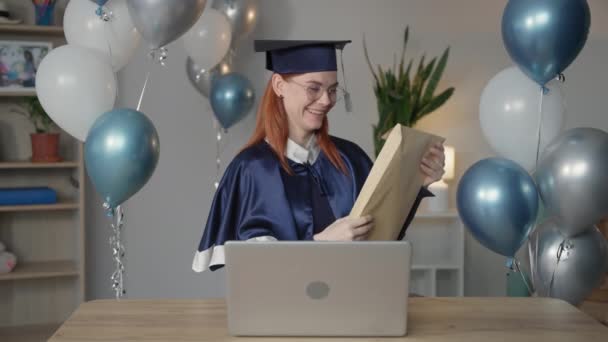 віртуальна церемонія випуску і скликання, радісна молода жінка в мантії радіє презентації диплома онлайн, використовуючи цифрові технології відеозв'язку на ноутбуці, сидячи вдома
 - Кадри, відео