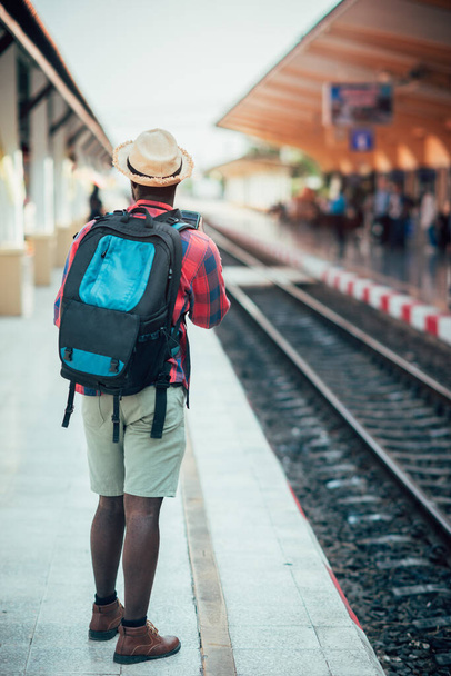Afrykański mężczyzna podróżnik z kapeluszem i plecakiem za pomocą smartfona i czeka na pociąg na stacji kolejowej.Adventure koncepcji podróży - Zdjęcie, obraz