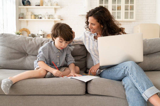 Schule zu Hause: Lächelnde Mutter hilft kleinem Sohn, während der Fernarbeit am Laptop in Notizbuch zu schreiben - Foto, Bild