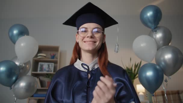encantadora mujer graduada en vestido académico es felizmente video chat en el ordenador portátil durante la ceremonia de graduación en línea mientras se sienta en casa durante la educación a distancia, - Imágenes, Vídeo