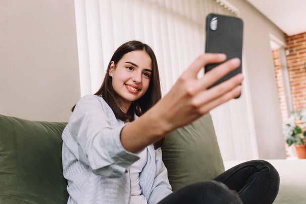 Pretty smiling teenager girl sitting at green sofa, posing a smile and taking a selfie with smartphone. High quality photo - Фото, изображение