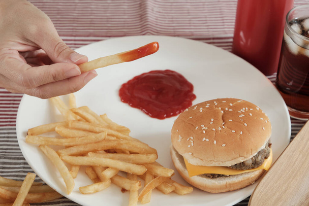 Tiro de close-up, foco seletivo: A mão do homem estava comendo frito francês, mergulhando ketchup em um prato branco em toalha de mesa vermelha com hambúrgueres e cola. Comer junk food ou fast food para o almoço não é saudável. - Foto, Imagem