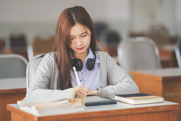Фотография молодой девушки-подростка азиатского колледжа в студенческой форме, которая учится и пишет на цифровом планшете в университетском классе - Фото, изображение