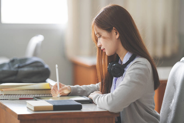 Foto d'archivio di una giovane adolescente studentessa asiatica in uniforme studentesca che studia e scrive su tablet digitale in una classe universitaria - Foto, immagini