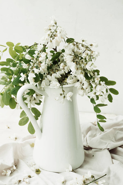 lush bouquet of white flowering acacia in white jug with bunch on light background. home decor with live plants. vertical content, selective focus - Фото, изображение