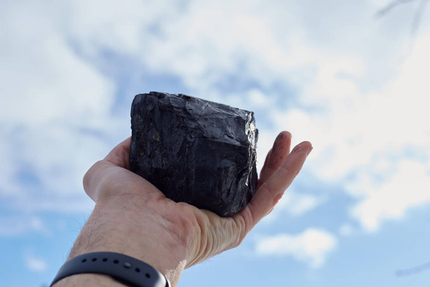Un uomo in mano tiene un carbone contro il cielo blu, inquinamento della natura. - Foto, immagini