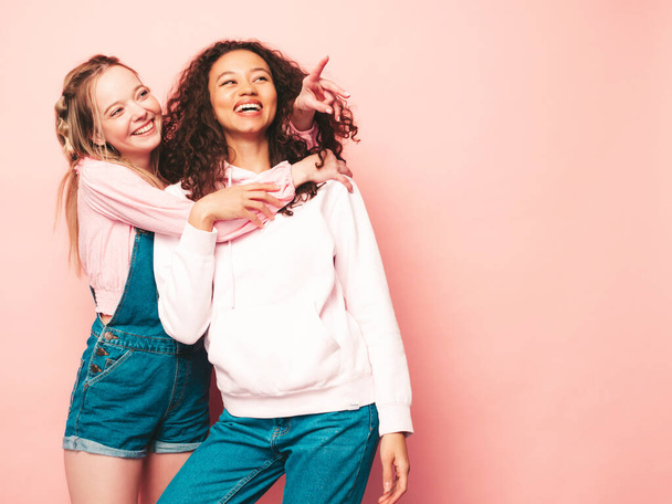Two young beautiful smiling international hipster female in trendy summer clothes. Sexy carefree women posing near pink wall in studio. Positive models having fun. Pointing on something  - Foto, immagini