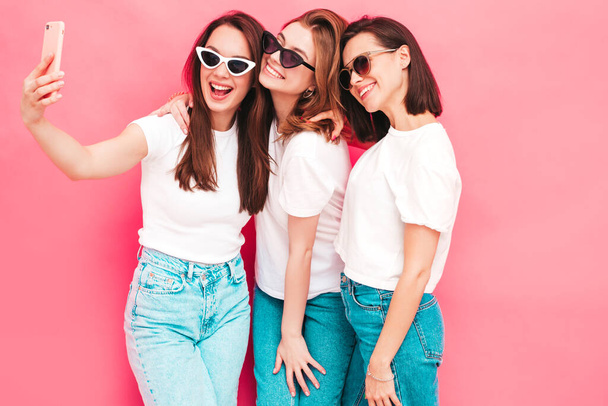 Three young beautiful smiling hipster female in trendy same summer white t-shirt and jeans clothes. Sexy carefree women posing near pink wall in studio. Positive models taking selfie photos - Valokuva, kuva