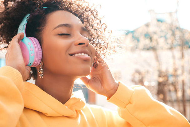 Beautiful black woman with afro curls hairstyle.Smiling model in yellow hoodie.Sexy carefree female enjoying listening music in wireless headphones.Posing on street background at sunset.Closed eyes - Фото, изображение