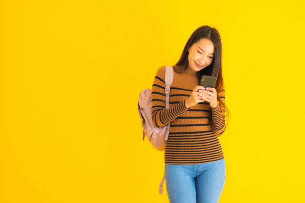 Портрет красива молода азіатка з рюкзаком використовує смартфон або мобільний телефон у багатьох діях на жовтому ізольованому фоні
 - Фото, зображення