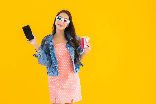 Portrait belle jeune femme asiatique profiter heureux avec téléphone pop-corn et regarder le film sur fond jaune isolé
 - Photo, image
